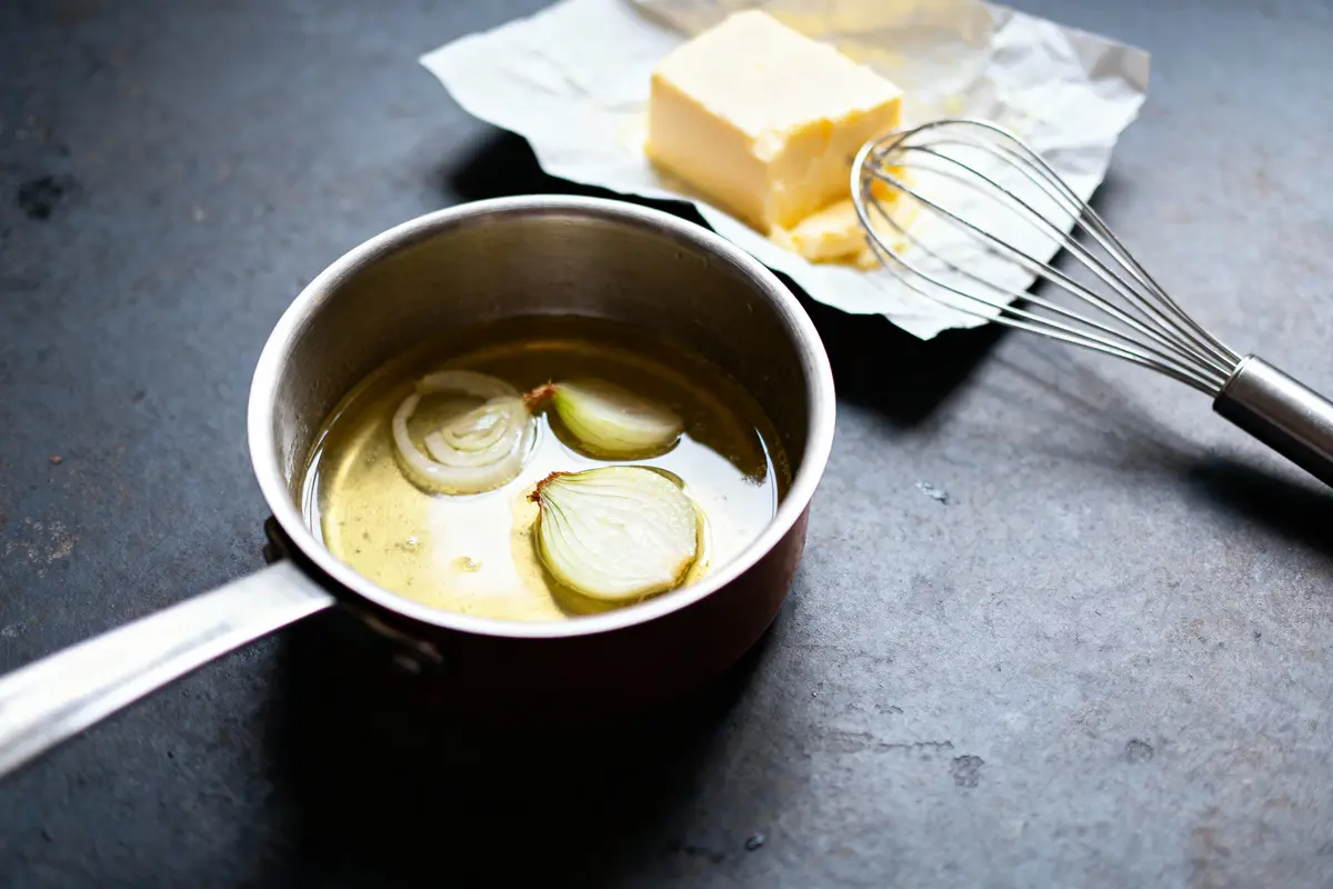 Casserole avec huile, rondelles d'oignon ; beurre sur papier, fouet en métal à côté.