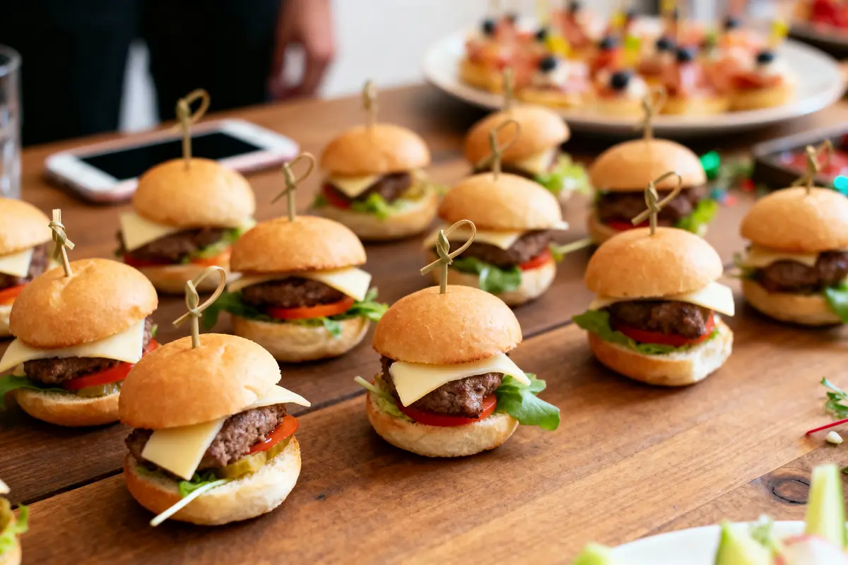 Petits burgers sur table en bois, garnis de fromage, tomates, laitue et brochettes décoratives. Téléphone en arrière-plan.