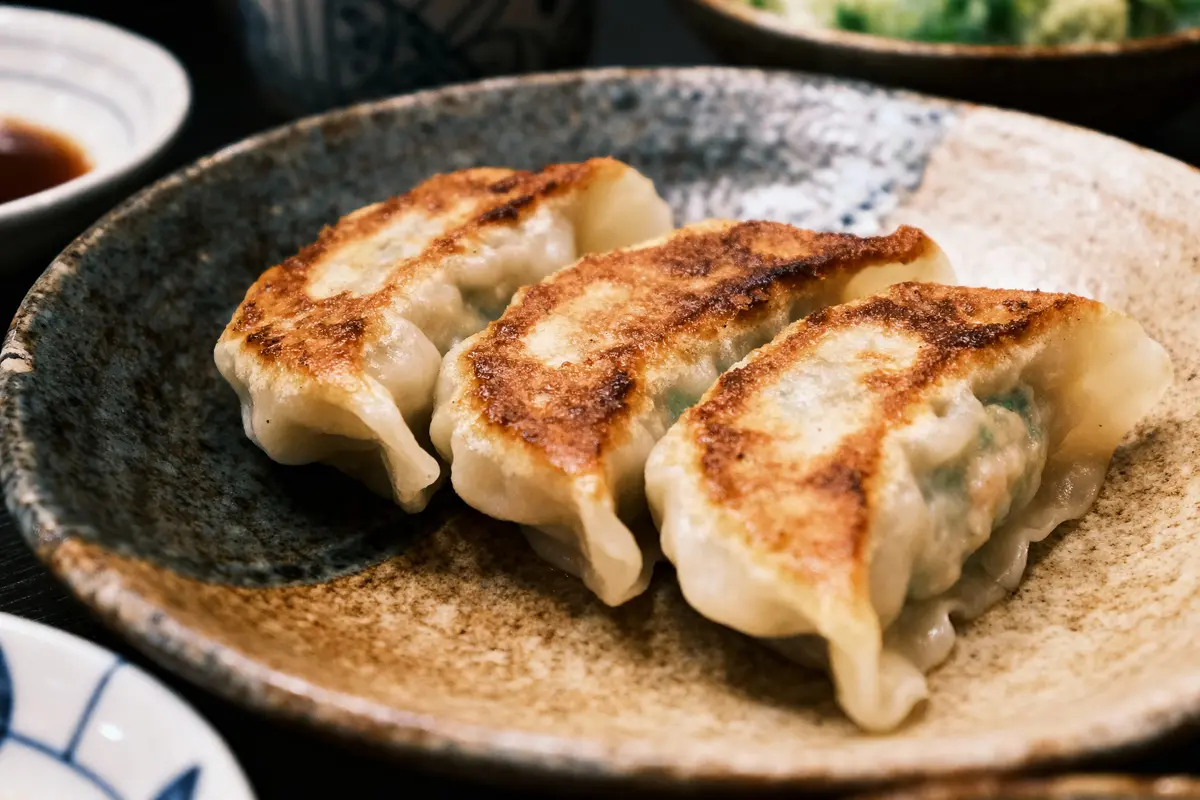 Trois gyozas grillés dans une assiette en céramique, accompagnés de sauce en arrière-plan.