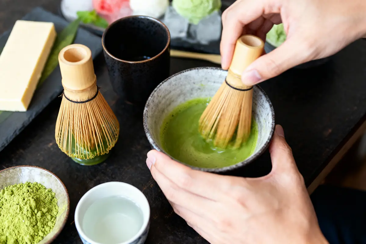 Mains préparant du thé matcha dans un bol avec fouet en bambou, poudre de matcha à côté.
