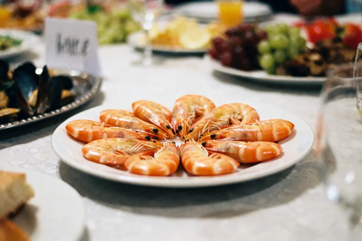 Assiette de crevettes disposées en cercle sur table, avec fruits et moules en arrière-plan.