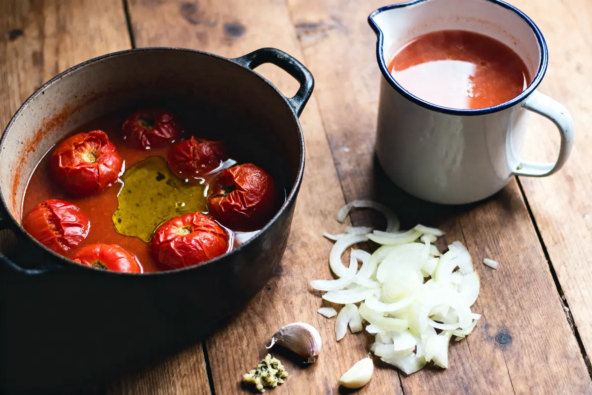 Cocotte avec tomates rôties, huile, casserole de sauce, oignons émincés, ail et herbes sur table en bois.