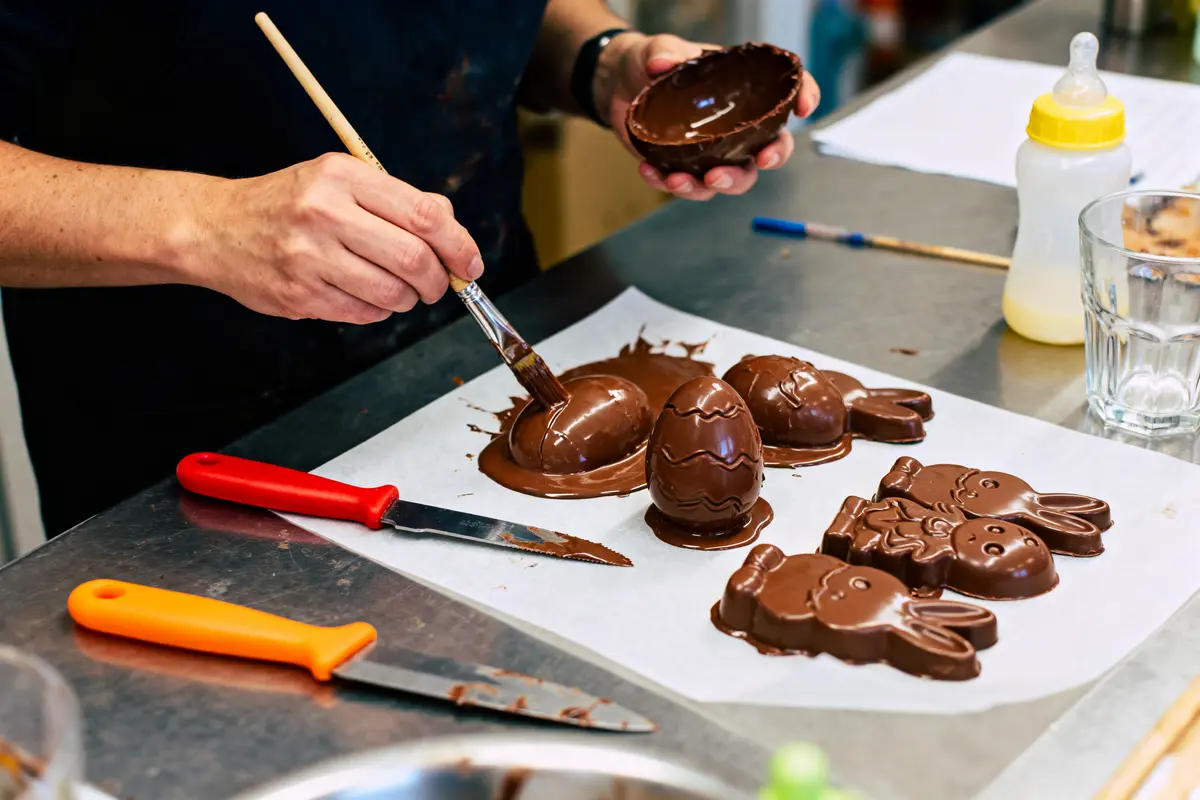 Personne décore des chocolats de Pâques maison sur un plan de travail avec pinceau et couteaux.
